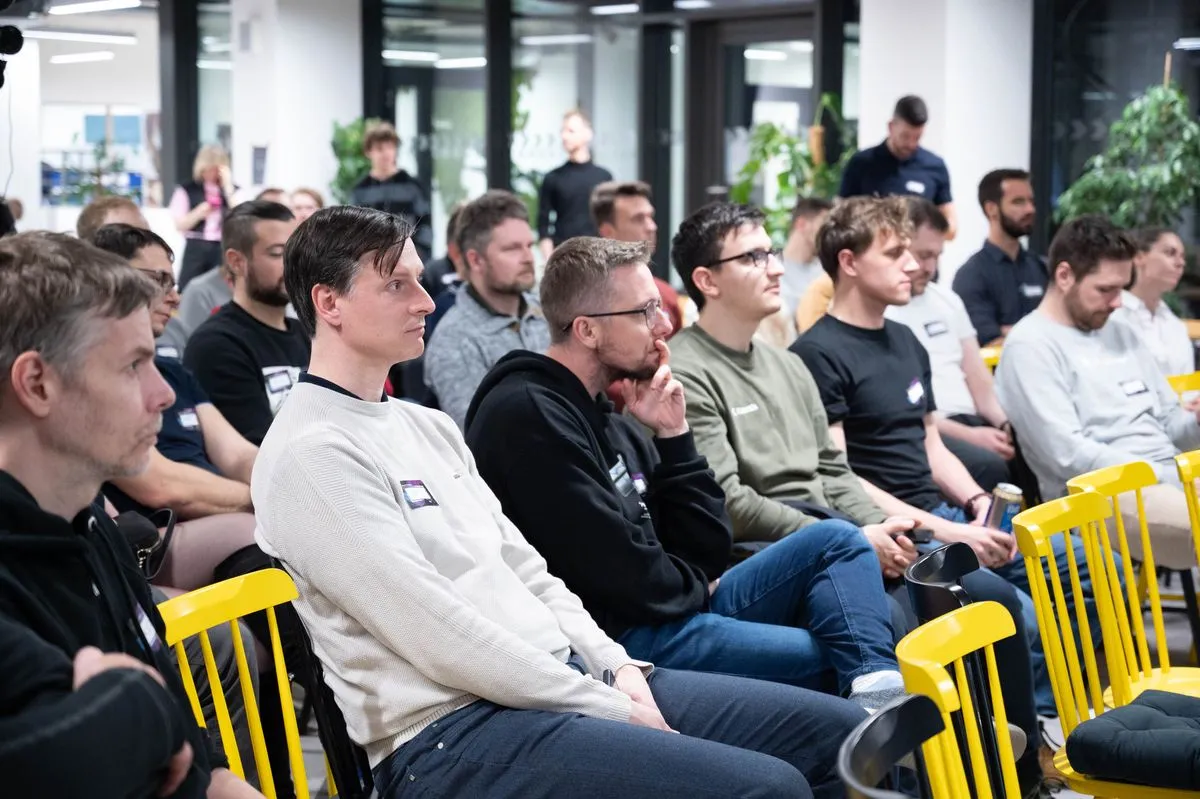 Rows of engineering leaders on yellow wooden chairs listening to a talk at an ELC meetup, more attendees standing by the glass office partitions behind them.