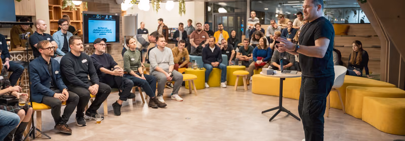 A speaker in a black t-shirt addressing about 40 seated engineering leaders at an ELC meetup, with a presentation screen behind them reading But there are challenges in bringing AI into your customer facing products.