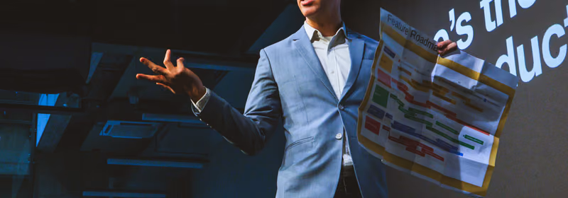 A speaker in a light blue blazer and round glasses wearing a headset microphone on a dark ELC stage, one hand open mid gesture, holding up a large printed sheet headed Feature Roadmap covered in coloured bars, with projected words on the wall behind him.