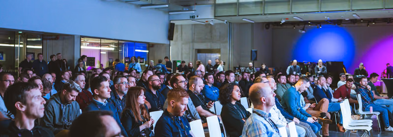 A large seated audience watching a talk at the ELC Conference in an industrial venue with exposed ceiling rigging, the stage behind them lit purple and blue.